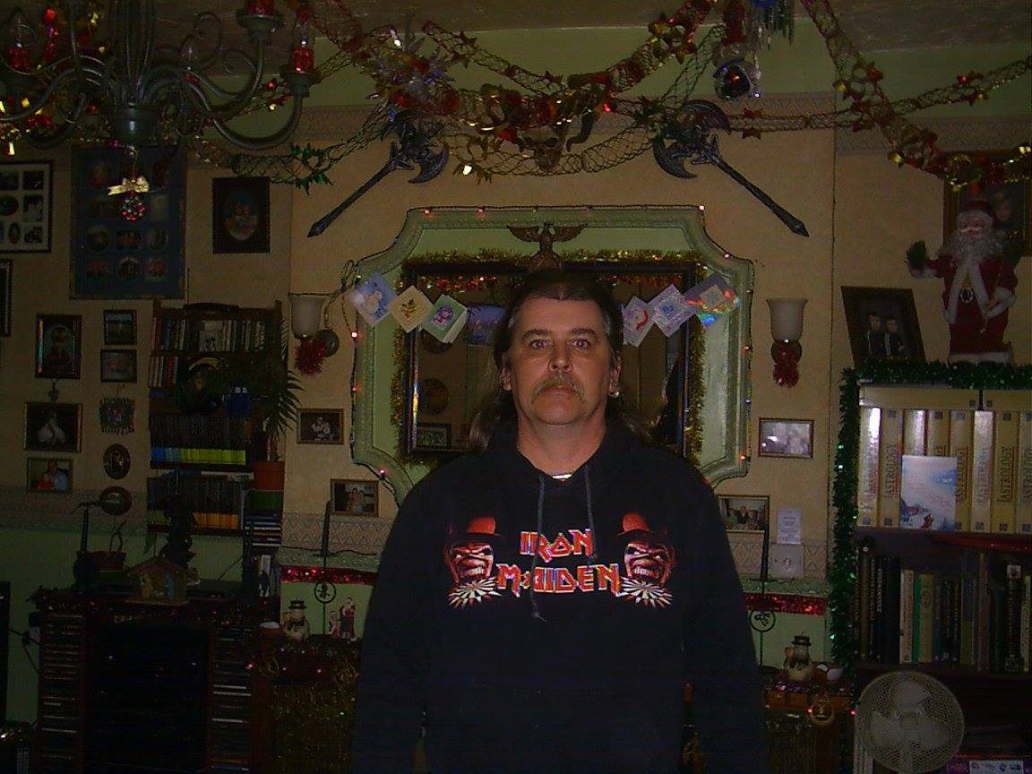 John standing in his front room in an Iron Maiden hoodie, tinsel and Christmas decorations around him.